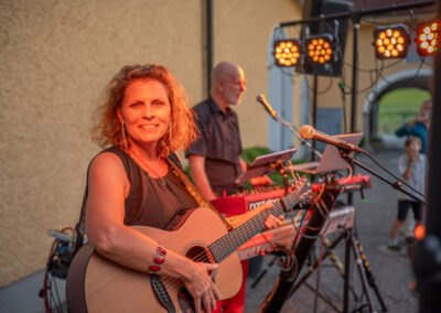 Lächelnde Hochzeitssängerin Nadja spielt Gitarre und Sänger Christian spielt Keyboard zum Tanz. Gelbe Scheinwerfer erzeugen Bühnenstimmung bei Hochzeitslocation Burnerhof Ansfelden.